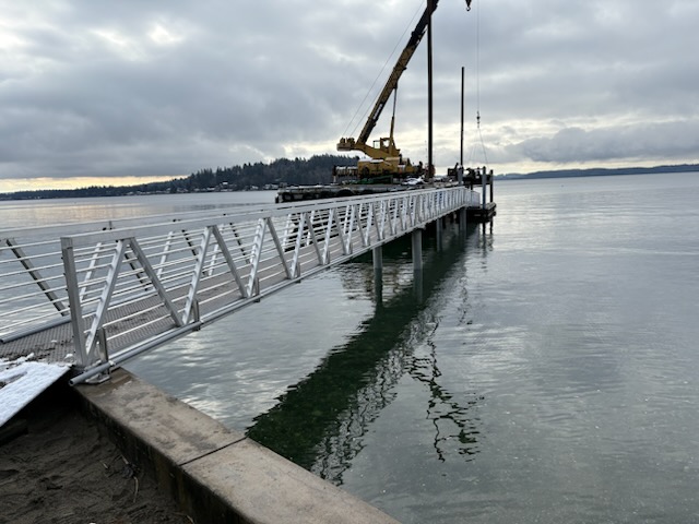 Crane installing piling on a dock accessed by metal gangway during marine construction in the Puget Sound region, with snow on the shoreline