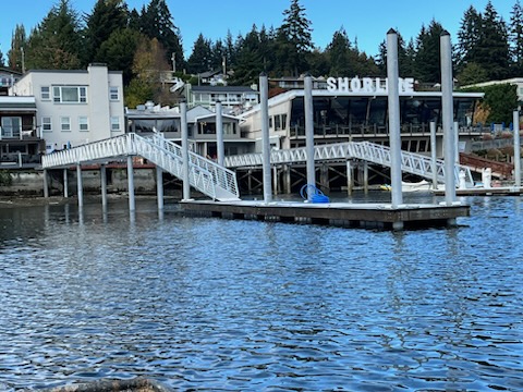 Floating dock with white metal gangway and tall pilings near a commercial waterfront building labeled SHORELINE in the Salish Sea region