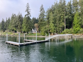 Floating dock with ramp extending into calm water, backed by evergreen forest and a shoreline residence in the Salish Sea region