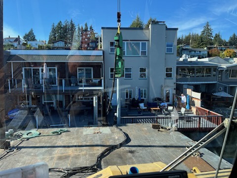 Crane operating on a marine construction barge near waterfront homes in the Salish Sea region, viewed from inside a control cabin